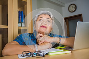 Attractive and tired middle aged Asian woman working at laptop computer desk confident daydreaming absent minded thinking about