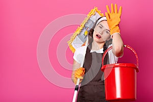 Attractive tired young female keeps hand on forehead, puts mop on shoulder. Bored housewife with opened mouth holds bucket and mop