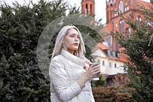 Attractive tranquil blond woman looking straight, drinking cup of coffee, wearing white warm jacket. Active tour weekend