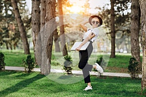 Attractive trendy happy hipster young woman with notebook in one hand in hat and sunglasses have fun in the park at the