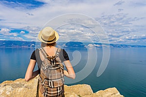 Attractive tween tourist girl in hat and backpack standing on cliff top and admiring beautiful landscape of blue sky and Baikal