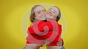 Attractive twin sisters hugging looking at camera posing with toothy smile at yellow background. Happy beautiful slim