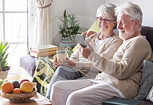 Attractive white-haired senior couple relaxing at home on the sofa looking together at smartphone in video call with family or