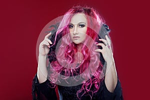 Attractive witch with red hair performs magic on a pink background. Halloween, horror theme.