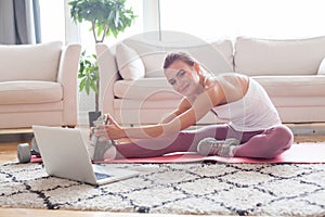 Attractive woman in activewear lying in front of laptop on pink mat while repeating exercise after instructor during online