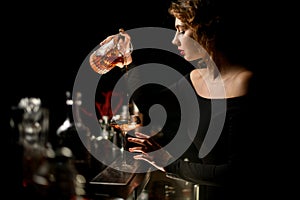 Attractive woman bartender in dark bar gently pours cocktail into wineglass.