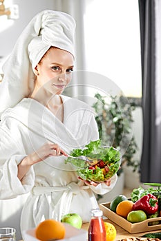 Attractive woman in bathrobe and towel eating salad, standing with lots of healthy fresh food in kitchen