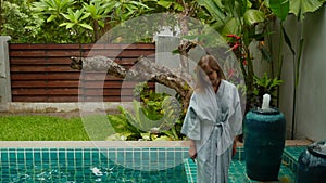 Attractive woman in bathrobe walks along edge of swimming pool in hotel room