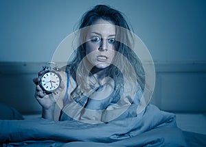 Attractive woman in bed showing alarm clock to camera feeling worried, stressed and sleepless