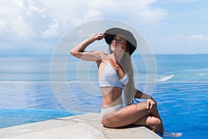 Attractive woman in bikini and with grey hat is relaxing in an infinity pool
