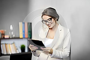 Attractive woman in black-framed glasses is smiling while reading or watching something on digital tablet