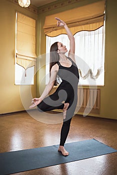 Attractive woman in black sportswear practicing yoga, doing Vrikshasana exercise, tree pose, exercising at home