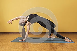 An attractive woman in black sportswear practicing yoga performs a kamatkarasana exercise in a room near the wall