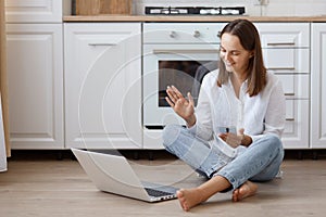 Attractive woman blogger wearing white t shirt and jeans sitting on floor in kitchen sitting in front of laptop with mobile phone