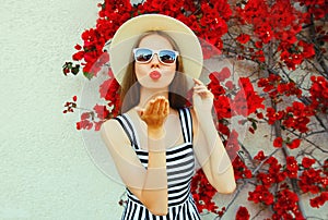 Attractive woman blowing red lips sending sweet air kiss wearing a summer straw hat over red flowers
