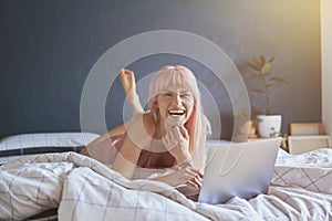 Attractive woman with bright pink laughs using modern laptop on bed by blue wall