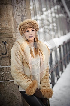 Attractive woman with brown fur cap and jacket enjoying the winter. Side view of fashionable blonde girl posing against bridge