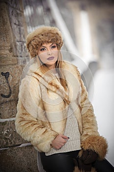 Attractive woman with brown fur cap and jacket enjoying the winter. Side view of fashionable blonde girl posing against bridge