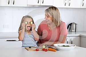 Attractive woman cooking together with her sweet beautiful blond little 6 or 7 years old daughter happy playful with cucumber as e