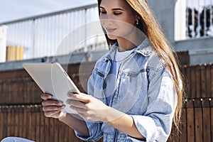 Attractive woman in denim clothes sits on park bench, holds smart tablet in her hands. Portrait of beautiful woman