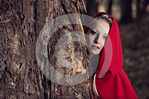 Attractive woman dressed a little red riding-hood hide behind a tree in dark forest