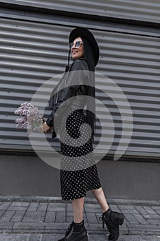 Attractive woman in fashion sunglasses in elegant hat in stylish black dress with bouquet of lilac poses near gray wall in street