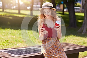 Attractive woman with foxy hair charges mobile phone on bench with solar panel, drinking coffee and checking email or types