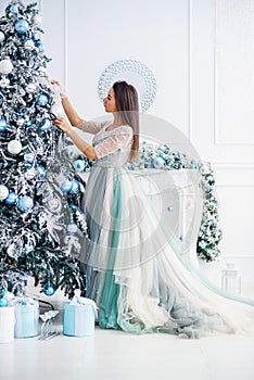 Attractive woman in a gentle evening dress decorates the Christmas tree in a cozy house. Happy winter holidays concept.