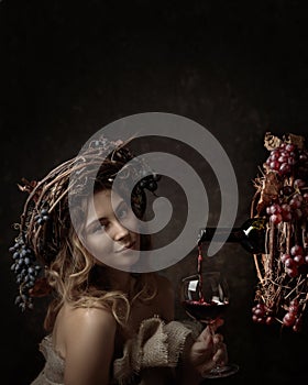 Attractive woman with glass of red wine in wine cellar . Vine wreath with blue grapes on a head. Red wine is poured from a bottle