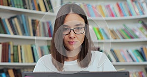 Attractive woman in glasses reading book and typing on computer. College student works on essay at the library.