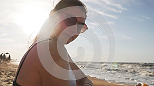 Attractive woman with healthy skin, applying sunscreen on her shoulder, hand while sitting on the beach near the sea