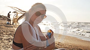 Attractive woman with healthy skin, applying sunscreen on her shoulder, hand while sitting on the beach near the sea