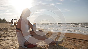 Attractive woman with healthy skin, applying sunscreen on her shoulder, hand while sitting on the beach near the sea
