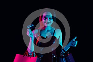 Attractive woman holding gift bags and bank credit card, looking at camera shows thumbs up smiling. Isolated on black