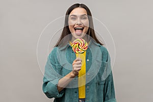 Attractive woman licking multicolor candy, wants to eat, looking at camera, showing tongue out.