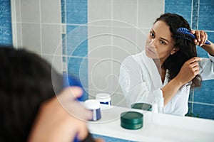 Attractive woman looking at her mirror reflection while combing hait in bathroom