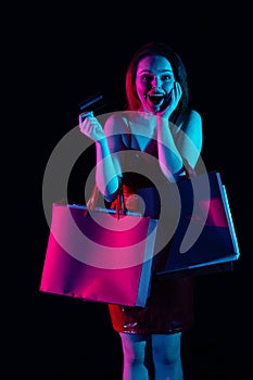Attractive woman with many shopping bags holding bank credit card with amazed smiling face expression. Isolated on black