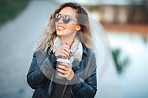 Attractive woman in mirrored sunglasses, a black leather jacket, drinking coffee on the waterfront river in the city in front the