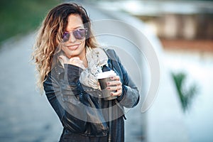 Attractive woman in mirrored sunglasses, a black leather jacket, drinking coffee on the waterfront river in the city in front the