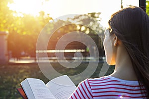 Attractive woman outdoors sitting on a bench reading a book in sunset. Back view