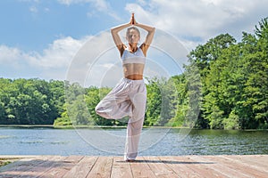 Attractive woman is practicing yoga, doing Vrksasana exercise, standing in tree pose near lake