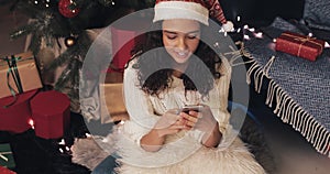 Attractive Woman in Santa`s hat Greeting Friends with Christmas Using Smartphone Typing Messages Sitting at Floor near