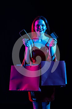 Attractive woman with shopping bags holding smartphone and credit card, looking at camera smiling. Isolated on black