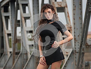 Attractive woman with short black dress and long leather boots standing on the rails with bridge in background. Fashion girl