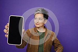 Attractive woman showing big smartphone blank screen? looking to screen and posing isolated over violet studio