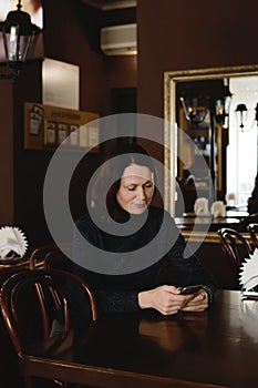 Attractive woman sits in a cafe with a beautiful interior and reads sms on her smartphone and smiles. The concept of gadgets,