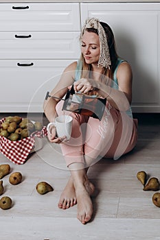 An attractive woman sits on the kitchen floor, pours tea into a mug. Pears are scattered around.