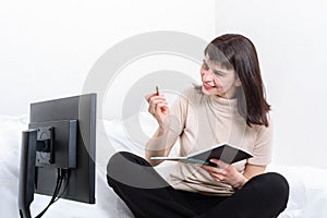 Attractive woman sitting on the couch holding a notebook and pen and looking intently at the computer monitor at home