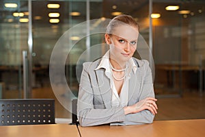 Attractive woman sitting at desk in meeting room looking friendly to the camera against blurred glass wall