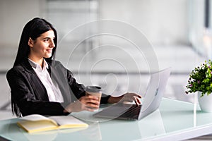 Attractive woman sitting at desk in office, working with laptop holding document, having takeaway coffee